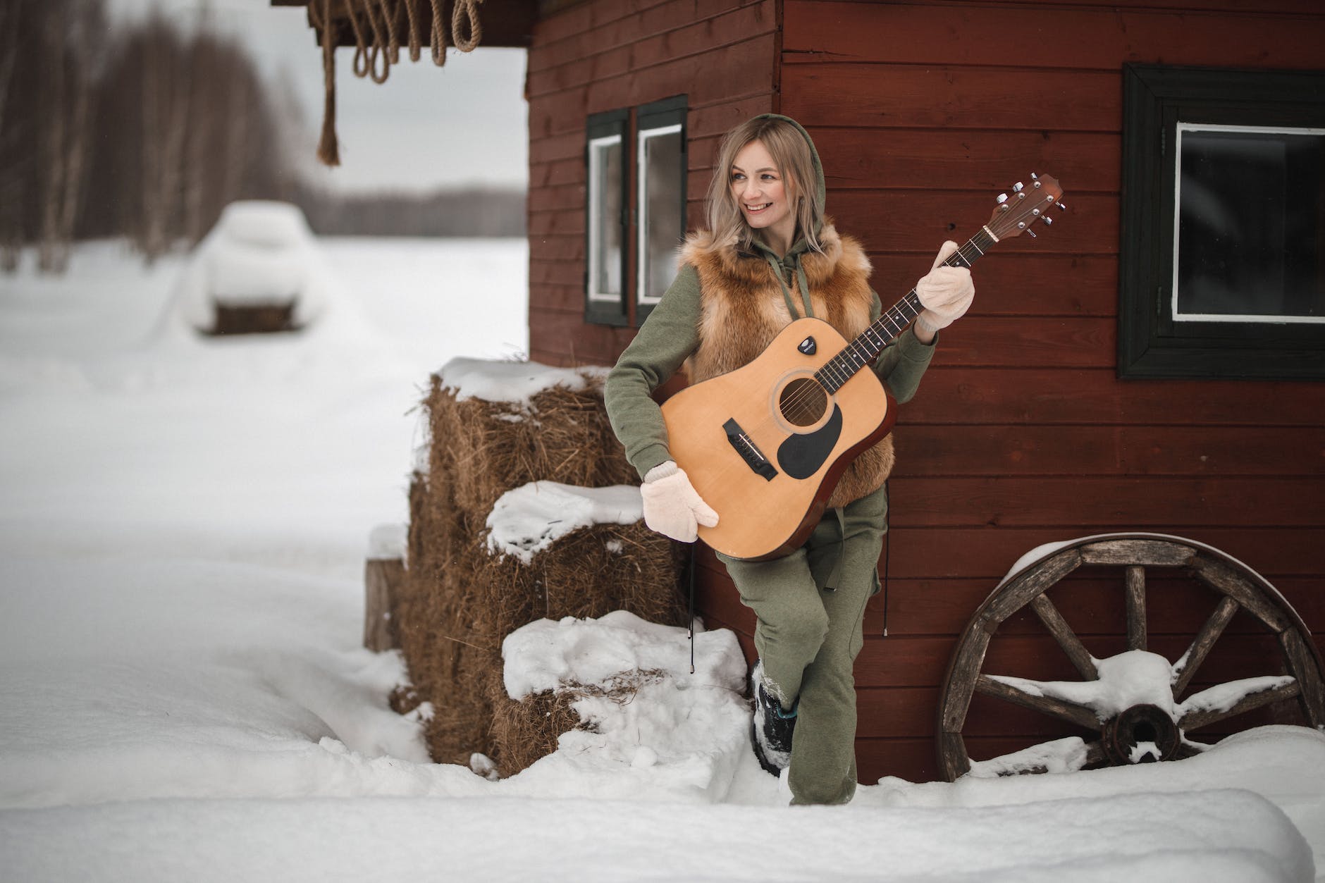 young cheerful woman standing by barn and holding classic guitar in winter scenery